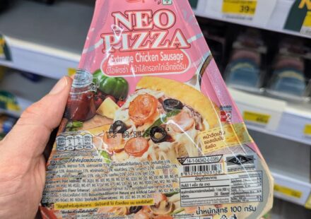 Alfredo NEO PIZZA Extreme Chicken Sausage: A Quick Hunger Fix That Falls Short on Crust and Cheese A hand holding a package of Alfredo NEO PIZZA Extreme Chicken Sausage inside a 7-Eleven store in Thailand.