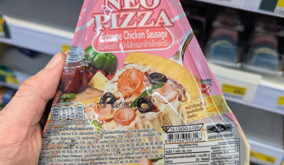 Alfredo NEO PIZZA Extreme Chicken Sausage: A Quick Hunger Fix That Falls Short on Crust and Cheese A hand holding a package of Alfredo NEO PIZZA Extreme Chicken Sausage inside a 7-Eleven store in Thailand.