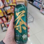 A hand holding a green 480ml bottle of Cha Jing Chinese Tea Osmanthus Flavour inside a 7-Eleven store in Thailand.