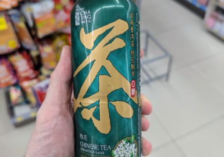A hand holding a green 480ml bottle of Cha Jing Chinese Tea Osmanthus Flavour inside a 7-Eleven store in Thailand.