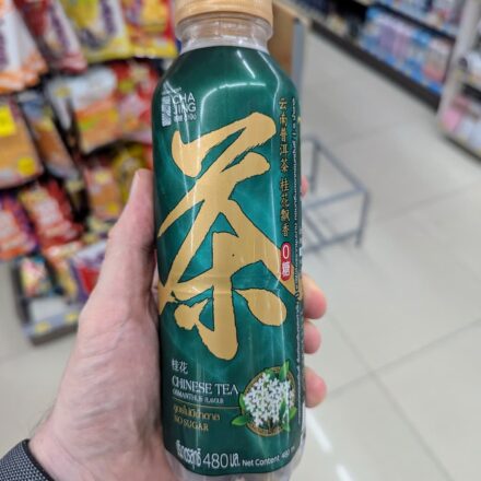 A hand holding a green 480ml bottle of Cha Jing Chinese Tea Osmanthus Flavour inside a 7-Eleven store in Thailand.