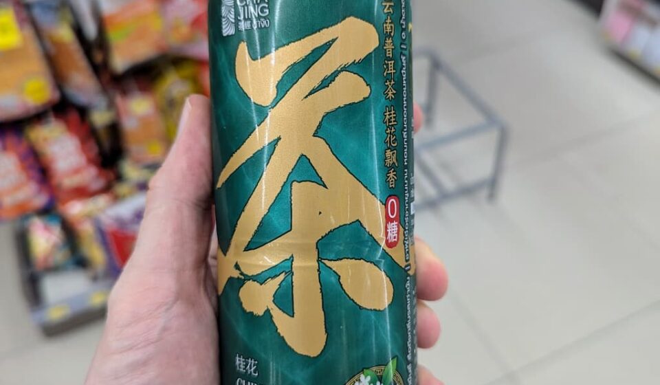 A hand holding a green 480ml bottle of Cha Jing Chinese Tea Osmanthus Flavour inside a 7-Eleven store in Thailand.