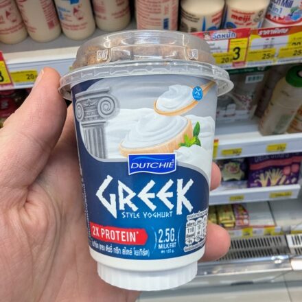 A hand holding a blue cup of Dutchie Greek Style Yoghurt with granola dome inside a 7-Eleven store in Thailand.