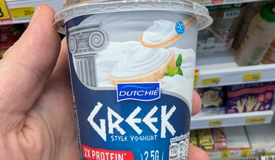 Dutchie Greek Style Yoghurt: Thick, Creamy, and High-Protein Value in Thailand A hand holding a blue cup of Dutchie Greek Style Yoghurt with granola dome inside a 7-Eleven store in Thailand.