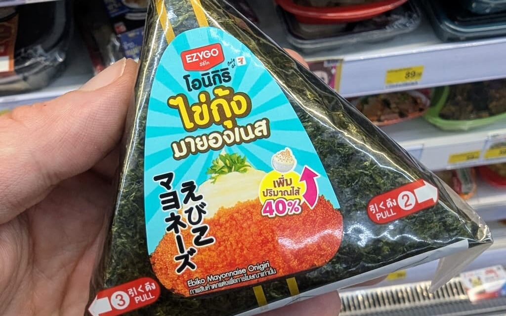 A hand holding a triangular package of Ezygo Ebiko Mayonnaise Onigiri inside a 7-Eleven store in Thailand.