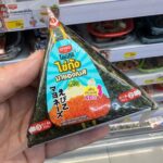 Alfredo NEO PIZZA Extreme Chicken Sausage: A Quick Hunger Fix That Falls Short on Crust and Cheese A hand holding a triangular package of Ezygo Ebiko Mayonnaise Onigiri inside a 7-Eleven store in Thailand.