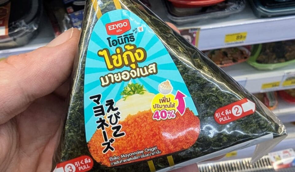 Ezygo Ebiko Mayonnaise Onigiri: A Crisp, Creamy Rice Snack That Hits the Spot Fresh A hand holding a triangular package of Ezygo Ebiko Mayonnaise Onigiri inside a 7-Eleven store in Thailand.