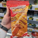 Ezygo Hot Dog Sausage with Bacon: A Fast, Soft-Bun Convenience Snack That Requires Heat A hand holding a red package of Ezygo Hot Dog Sausage with Bacon inside a 7-Eleven store in Thailand.