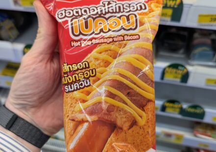 Ezygo Hot Dog Sausage with Bacon: A Fast, Soft-Bun Convenience Snack That Requires Heat A hand holding a red package of Ezygo Hot Dog Sausage with Bacon inside a 7-Eleven store in Thailand.