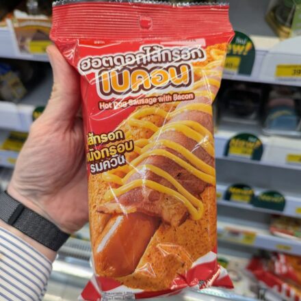 A hand holding a red package of Ezygo Hot Dog Sausage with Bacon inside a 7-Eleven store in Thailand.