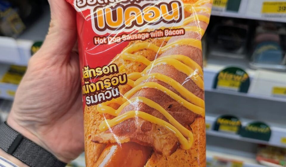 Ezygo Hot Dog Sausage with Bacon: A Fast, Soft-Bun Convenience Snack That Requires Heat A hand holding a red package of Ezygo Hot Dog Sausage with Bacon inside a 7-Eleven store in Thailand.