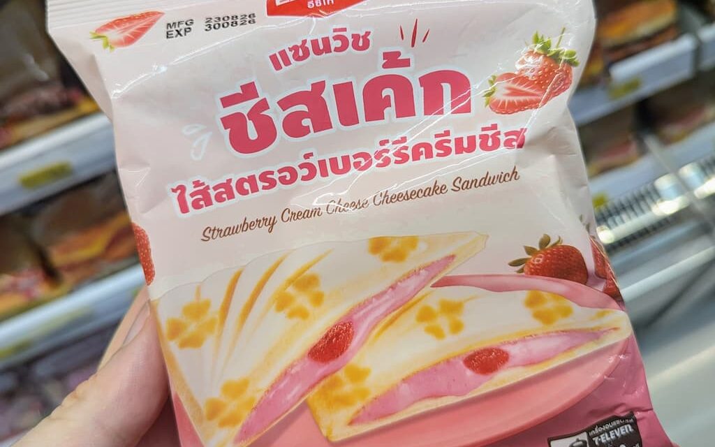 A hand holding a pastel pink package of Ezygo Strawberry Cream Cheese Cheesecake Sandwich inside a 7-Eleven store in Thailand.