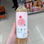 Ezygo Hot Dog Sausage with Bacon: A Fast, Soft-Bun Convenience Snack That Requires Heat A hand holding a slim 450ml bottle of HEYTEA Peach Cranberry Green Tea inside a 7-Eleven store in Thailand.