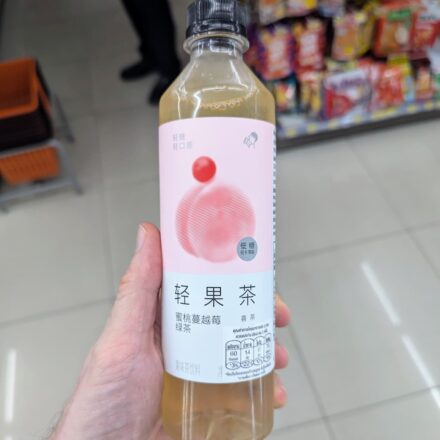A hand holding a slim 450ml bottle of HEYTEA Peach Cranberry Green Tea inside a 7-Eleven store in Thailand.