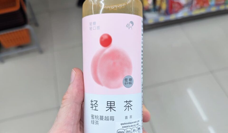 HEYTEA Peach Cranberry Green Tea: A Refreshing Chinese Fruit Tea Picked on Recommendation A hand holding a slim 450ml bottle of HEYTEA Peach Cranberry Green Tea inside a 7-Eleven store in Thailand.