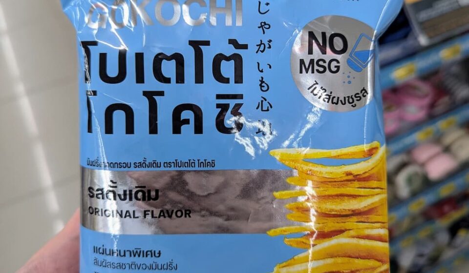 Koikeya Potato Kochi Original Flavor: Thick-Cut, Pure Potato Crunch with Zero MSG A hand holding a light blue bag of Koikeya Potato Kochi Original Flavor chips inside a 7-Eleven store in Thailand.