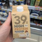 Magnum Ice Cream Promo at 7-Eleven Thailand: 39 THB Deal & Delivery Guide A hand holding a beige 400ml carton of Mülk+ 39g High Protein Chocolate Lactose Free milk in front of a 7-Eleven display in Thailand.