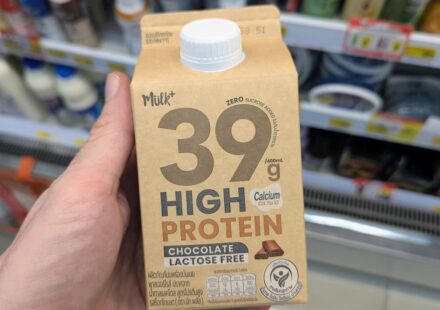 Mülk+ 39g High Protein Chocolate Milk (Lactose Free): Massive Protein Power with Great Cocoa Flavor A hand holding a beige 400ml carton of Mülk+ 39g High Protein Chocolate Lactose Free milk in front of a 7-Eleven display in Thailand.