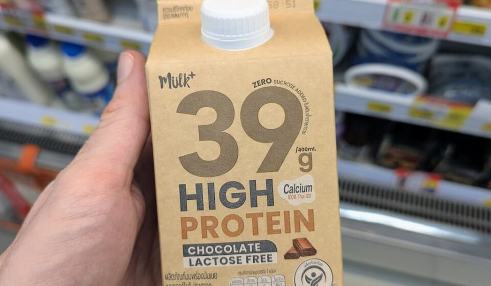 Mülk+ 39g High Protein Chocolate Milk (Lactose Free): Massive Protein Power with Great Cocoa Flavor A hand holding a beige 400ml carton of Mülk+ 39g High Protein Chocolate Lactose Free milk in front of a 7-Eleven display in Thailand.