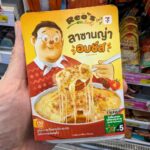 A hand holding a yellow box of Reo's Deli Baked Cheese Lasagna inside a 7-Eleven store in Thailand.
