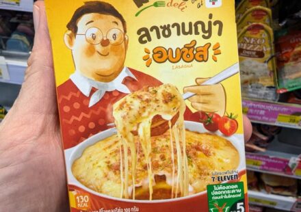 A hand holding a yellow box of Reo's Deli Baked Cheese Lasagna inside a 7-Eleven store in Thailand.