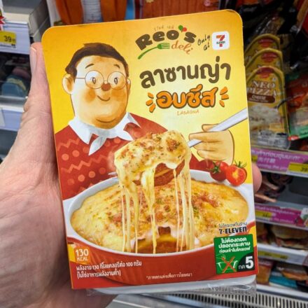 A hand holding a yellow box of Reo's Deli Baked Cheese Lasagna inside a 7-Eleven store in Thailand.