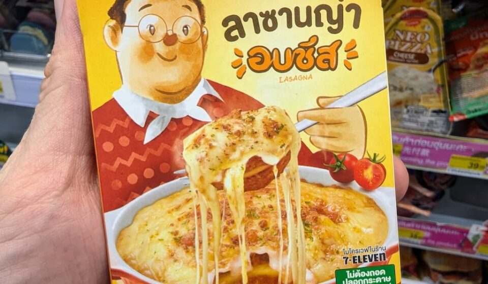 A hand holding a yellow box of Reo's Deli Baked Cheese Lasagna inside a 7-Eleven store in Thailand.