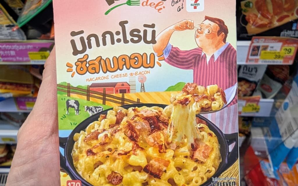 A hand holding a pink box of Reo's Deli Macaroni Cheese & Bacon inside a 7-Eleven store in Thailand.