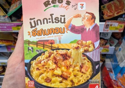 Reo’s Deli Macaroni Cheese & Bacon: Creamy Comfort Food with a Slightly Sweet Thai Twist A hand holding a pink box of Reo's Deli Macaroni Cheese & Bacon inside a 7-Eleven store in Thailand.