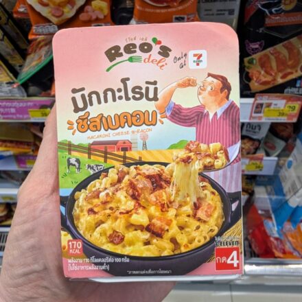 A hand holding a pink box of Reo's Deli Macaroni Cheese & Bacon inside a 7-Eleven store in Thailand.