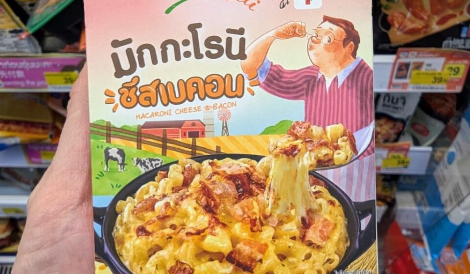 Reo’s Deli Macaroni Cheese & Bacon: Creamy Comfort Food with a Slightly Sweet Thai Twist A hand holding a pink box of Reo's Deli Macaroni Cheese & Bacon inside a 7-Eleven store in Thailand.