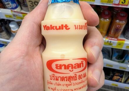 A hand holding an iconic 80ml bottle of Yakult Probiotic Cultured Milk in front of a 7-Eleven dairy display in Thailand.